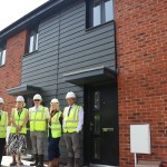 Five people in hi-vis vests and hard hats stand outside a new red brick and dark-clad house at the Garden Village development