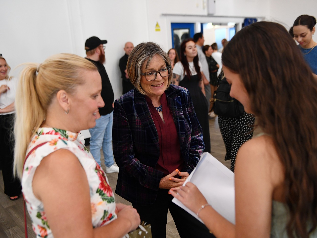 Education Minister Anna Brychan smiling as she talks to a student holding a results envelope and her parent in a school hall