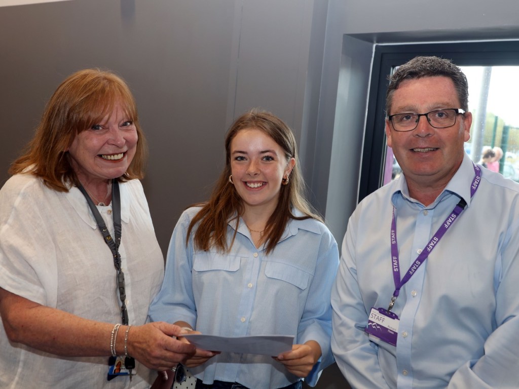 A smiling teenage girl holding her results sheet between two smiling members of school staff