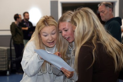 Three teenage girls huddled together reading a GCSE results slip in a school hall, one with a look of surprise