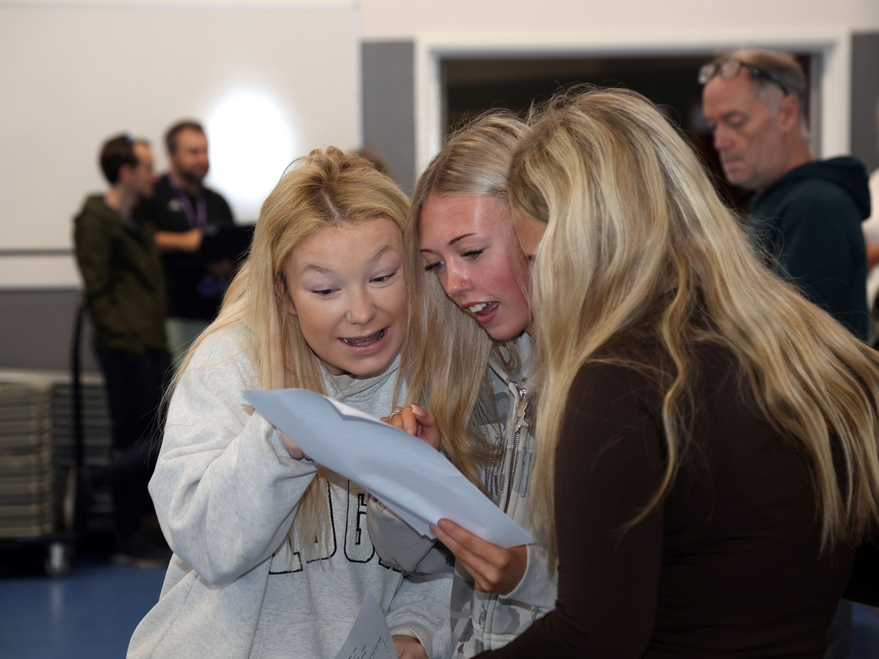 Three teenage girls huddled together reading a GCSE results slip in a school hall, one with a look of surprise