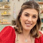 Georgie Grasso smiling in a red dress and apron in the Bake Off tent, shelves of baking equipment behind her