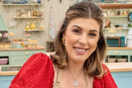 Georgie Grasso smiling in a red dress and apron in the Bake Off tent, shelves of baking equipment behind her