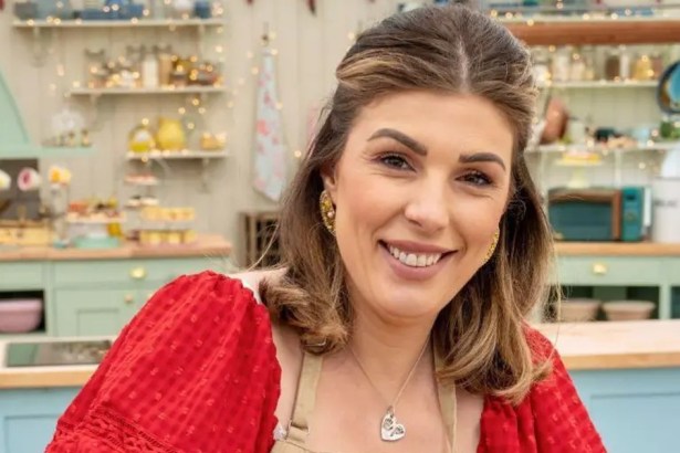 Georgie Grasso smiling in a red dress and apron in the Bake Off tent, shelves of baking equipment behind her