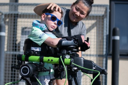 A boy in blue sunglasses stands in a Trexo robotic exoskeleton while a smiling physiotherapist supports him