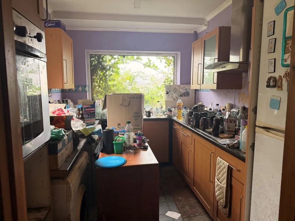 A kitchen with worktops and units still loaded with belongings, boxes and bottles, and damp on the wall