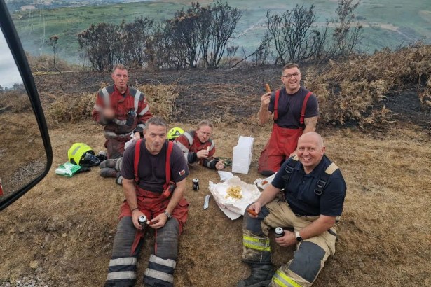 Five Mid and West Wales firefighters sitting on a scorched hillside eating sausage and chips, with burnt gorse behind them and green hills beyond