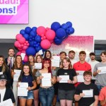 A large group of students holding results slips in front of balloons and a Congratulations screen at Gower College Swansea