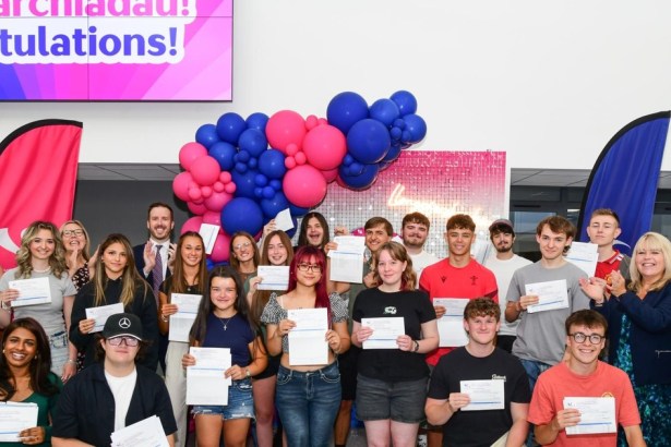 A large group of students holding results slips in front of balloons and a Congratulations screen at Gower College Swansea