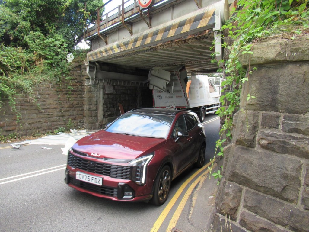 A red car passing slowly under the Gowerton railway bridge beside the wedged lorry with debris on the road
