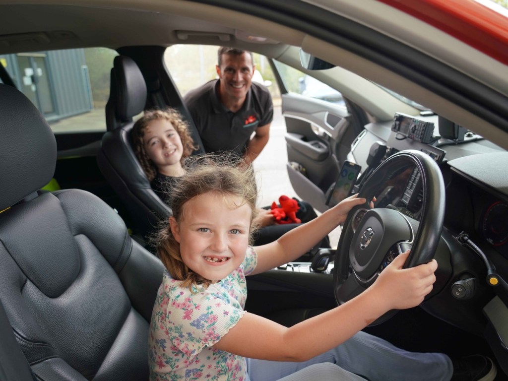 Gracie sitting behind the wheel of a Wales Air Ambulance rapid response vehicle with her brother Chester and a critical care practitioner