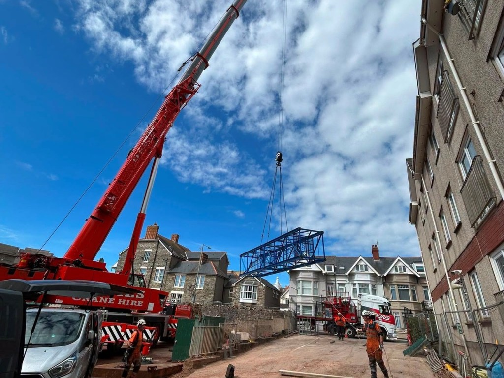 Two workers in orange overalls watch as a mobile crane lifts a tower crane section over houses beside the Grand Pavilion in Porthcawl