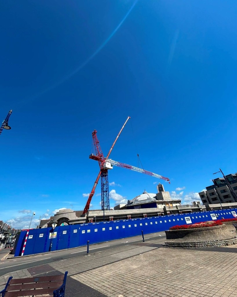 The jib of a red and white tower crane extends over the clock tower and dome of the Grand Pavilion in Porthcawl against a clear blue sky