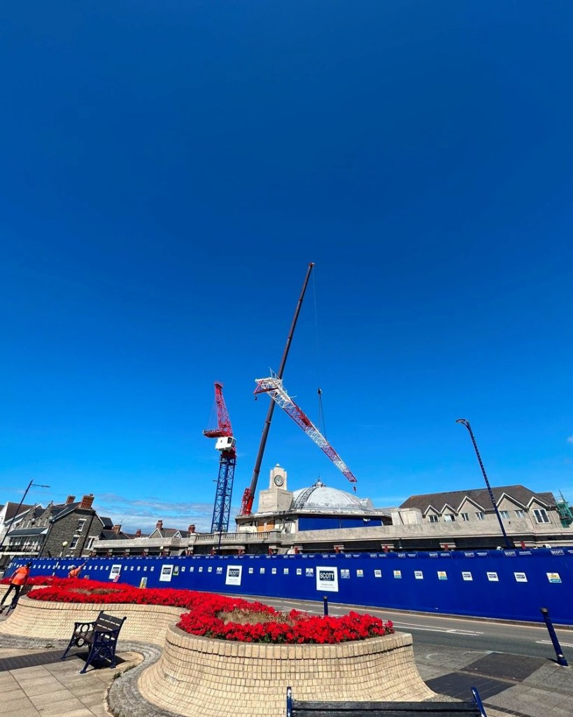 A tower crane and a mobile crane stand above the domed roof and clock tower of the Grand Pavilion in Porthcawl behind blue hoardings