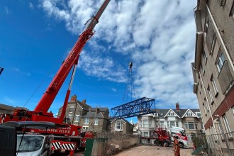 A red mobile crane lifts a blue lattice tower crane section over Victorian terraced houses under a blue sky in Porthcawl
