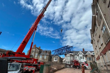 A red mobile crane lifts a blue lattice tower crane section over Victorian terraced houses under a blue sky in Porthcawl