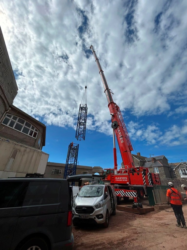 A blue lattice tower crane section hangs from a red mobile crane above the partly built tower on the Grand Pavilion site in Porthcawl