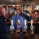 A smiling barman rings a handbell behind a pub bar lined with Greene King IPA and Abbot Ale hand pumps, as two customers laugh in the foreground