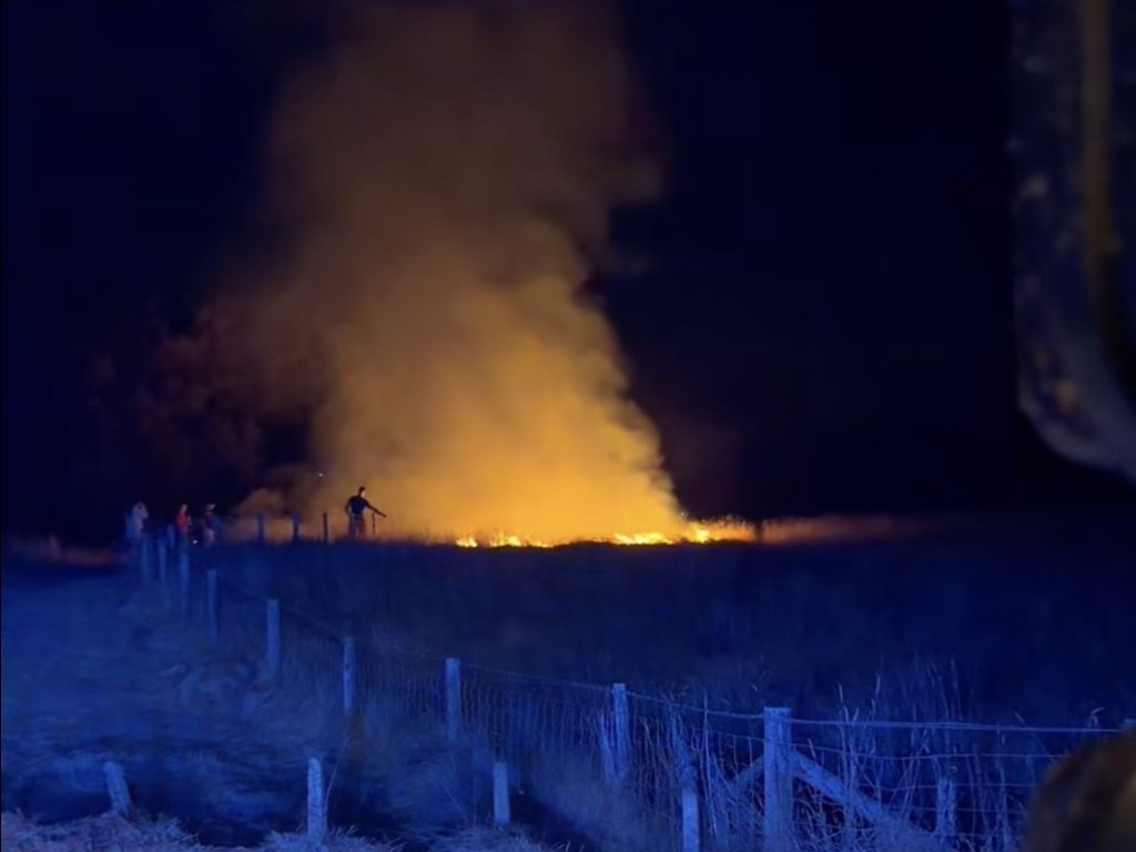 A wide night-time view of smoke and flames on Gwaun Cae Gurwen common, with a fence line in the blue-lit foreground