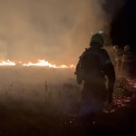 Firefighters silhouetted against a line of flames and smoke on Gwaun Cae Gurwen common at night, with embers glowing on the ground