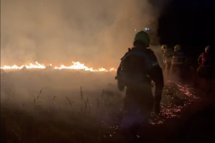 Firefighters silhouetted against a line of flames and smoke on Gwaun Cae Gurwen common at night, with embers glowing on the ground