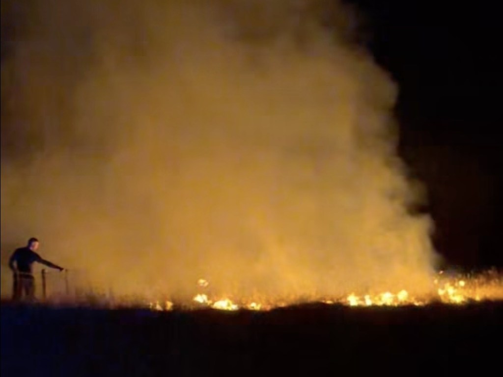 A large plume of smoke lit orange by flames on Gwaun Cae Gurwen common at night, with a person standing at a fence to the left