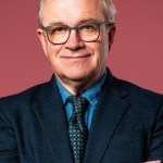 Harry Enfield in glasses and a dark suit with arms folded against a pink background