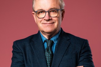 Harry Enfield in glasses and a dark suit with arms folded against a pink background