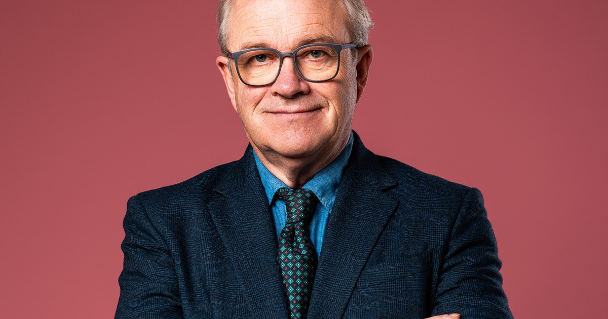 Harry Enfield in glasses and a dark suit with arms folded against a pink background