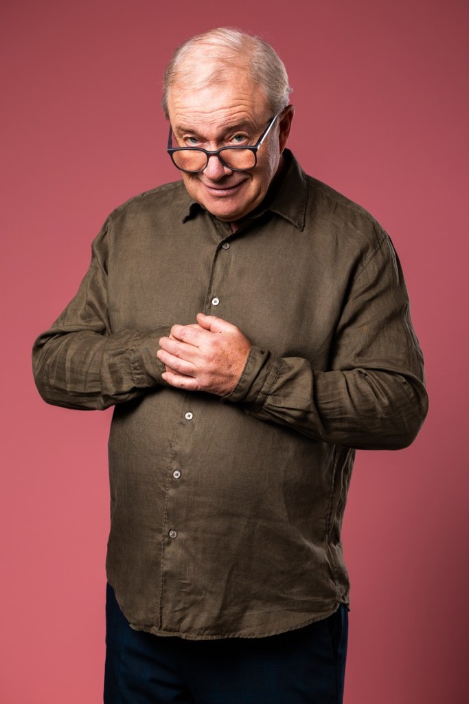 Harry Enfield peering over the top of his glasses with hands clasped, against a pink background