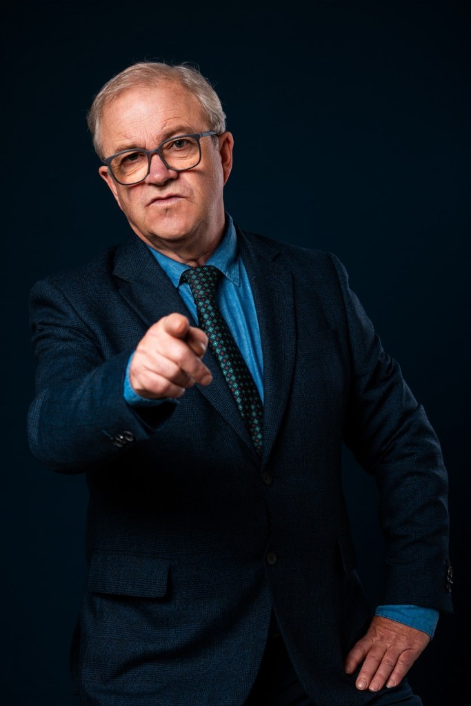 Harry Enfield pointing at the camera with a stern expression in a dark suit