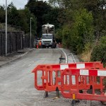 A closed road in Pontyberem with new kerbs laid along a stone retaining wall, red barriers in the foreground and a tarmac lorry and workers in orange further along