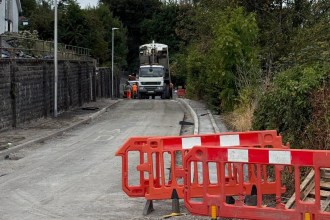 A closed road in Pontyberem with new kerbs laid along a stone retaining wall, red barriers in the foreground and a tarmac lorry and workers in orange further along