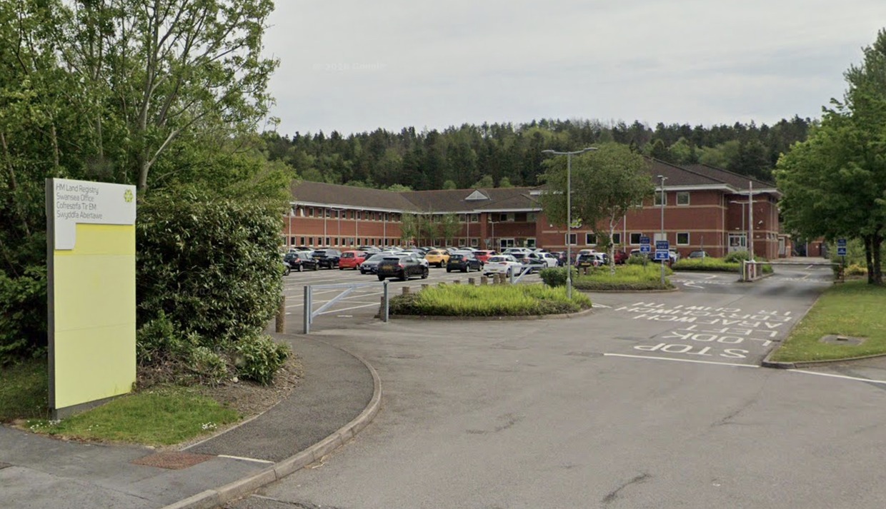 The entrance to HM Land Registry's Swansea office, with a bilingual sign beside the access road and the red-brick office building behind a car park