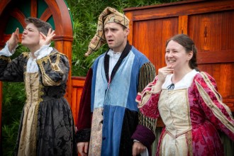 Three actors in Elizabethan costume on Illyria's outdoor stage, one in a patchwork jester's outfit and cap