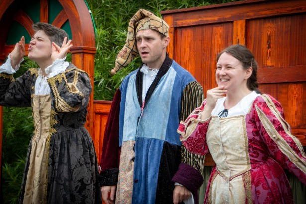 Three actors in Elizabethan costume on Illyria's outdoor stage, one in a patchwork jester's outfit and cap