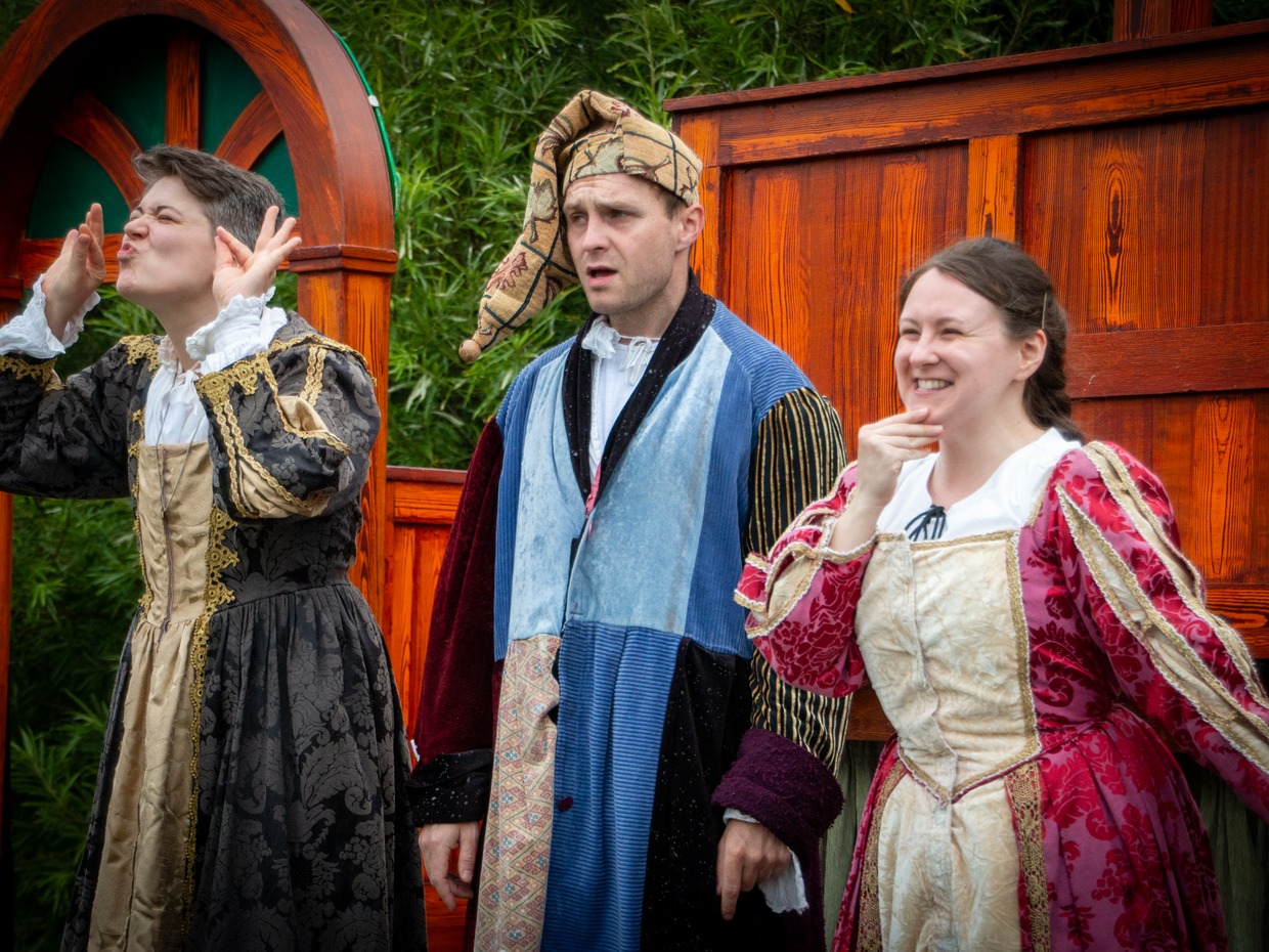 Three actors in Elizabethan costume on Illyria's outdoor stage, one in a patchwork jester's outfit and cap