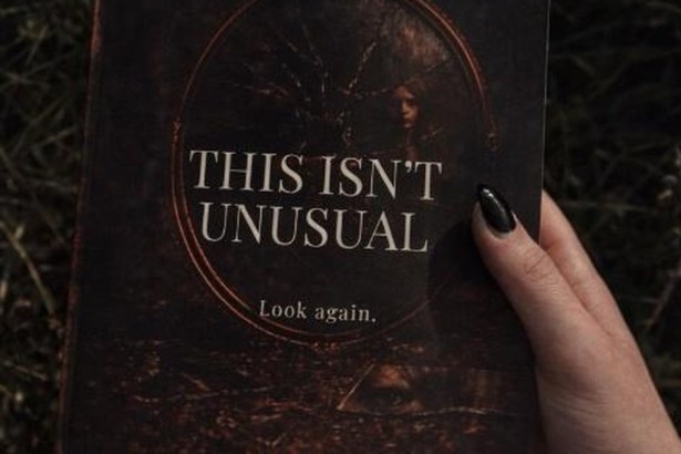 A hand holding a copy of the psychological horror collection This Isn't Unusual by A. Sjoberg against dark undergrowth