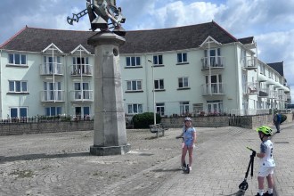 Two children on scooters beside a sculpture mounted on a stone column at Swansea Marina