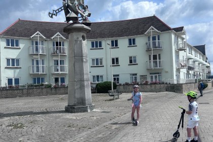 Two children on scooters beside a sculpture mounted on a stone column at Swansea Marina
