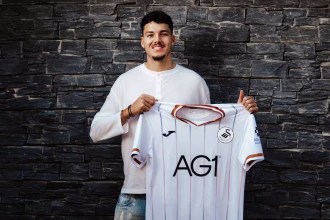 Jenson Seelt smiling and holding up a white Swansea City home shirt in front of a dark stone wall