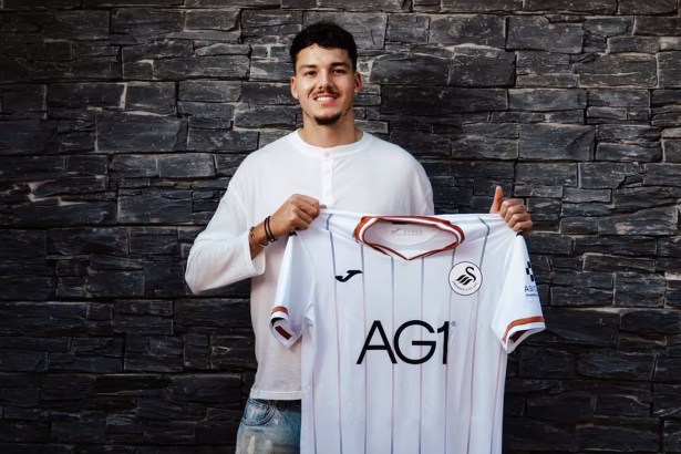 Jenson Seelt smiling and holding up a white Swansea City home shirt in front of a dark stone wall