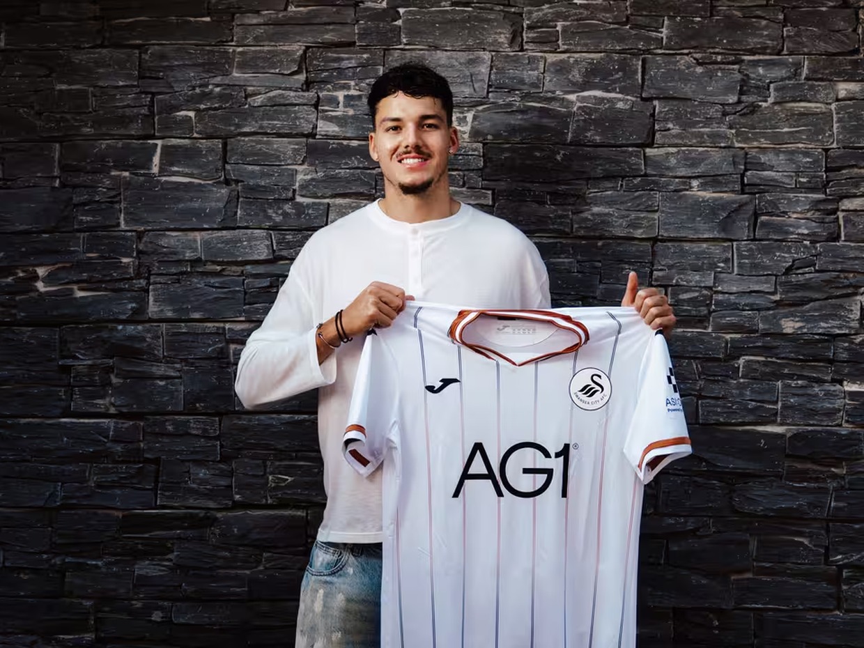 Jenson Seelt smiling and holding up a white Swansea City home shirt in front of a dark stone wall