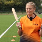 Katy Goss in an orange My Rounders shirt holding a rounders bat and ball on an artificial pitch
