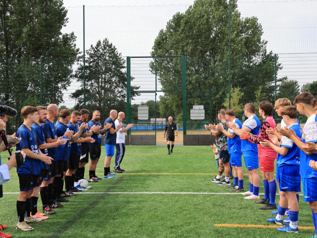 Two lines of footballers in black-and-blue and blue-and-white kits applaud as referee Ken Wright walks out between them on a 3G pitch