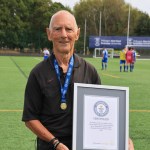 Ken Wright in a black referee's shirt and medal holding his framed Guinness World Records certificate on a 3G pitch, with players warming up behind him