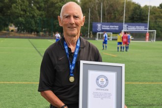 Ken Wright in a black referee's shirt and medal holding his framed Guinness World Records certificate on a 3G pitch, with players warming up behind him