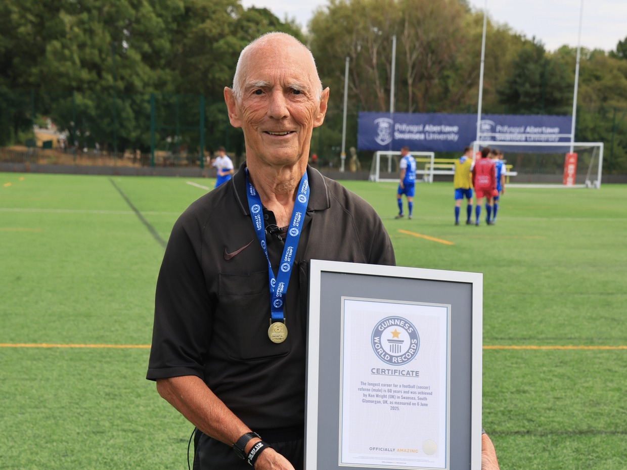 Ken Wright in a black referee's shirt and medal holding his framed Guinness World Records certificate on a 3G pitch, with players warming up behind him