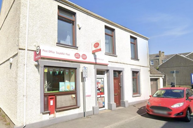 The white-fronted Kidwelly Post Office on Station Road, with bilingual Post Office signage, a red wall postbox and Welsh flag bunting in the window