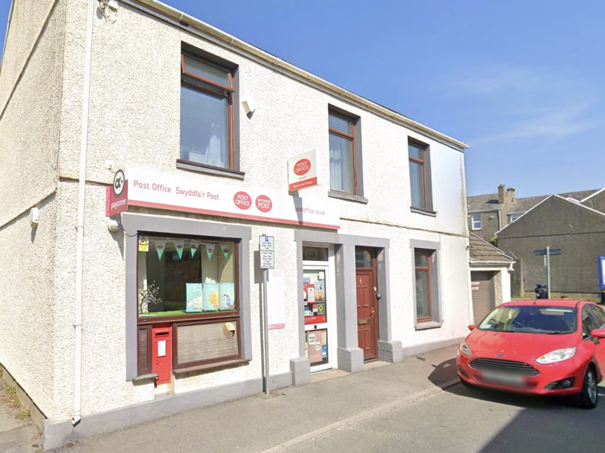 The white-fronted Kidwelly Post Office on Station Road, with bilingual Post Office signage, a red wall postbox and Welsh flag bunting in the window